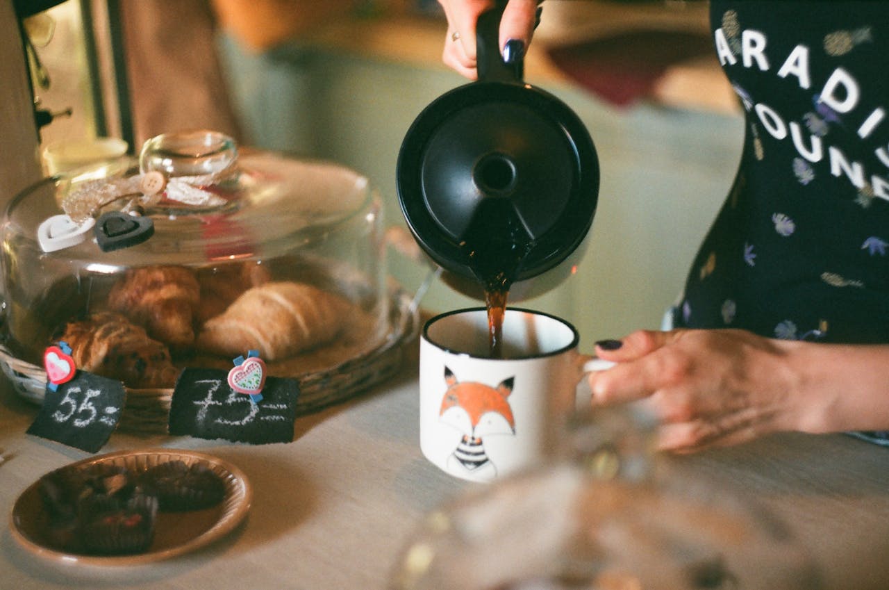 A person pours coffee into a fox-themed mug next to pastries under a glass dome in a cozy setting.